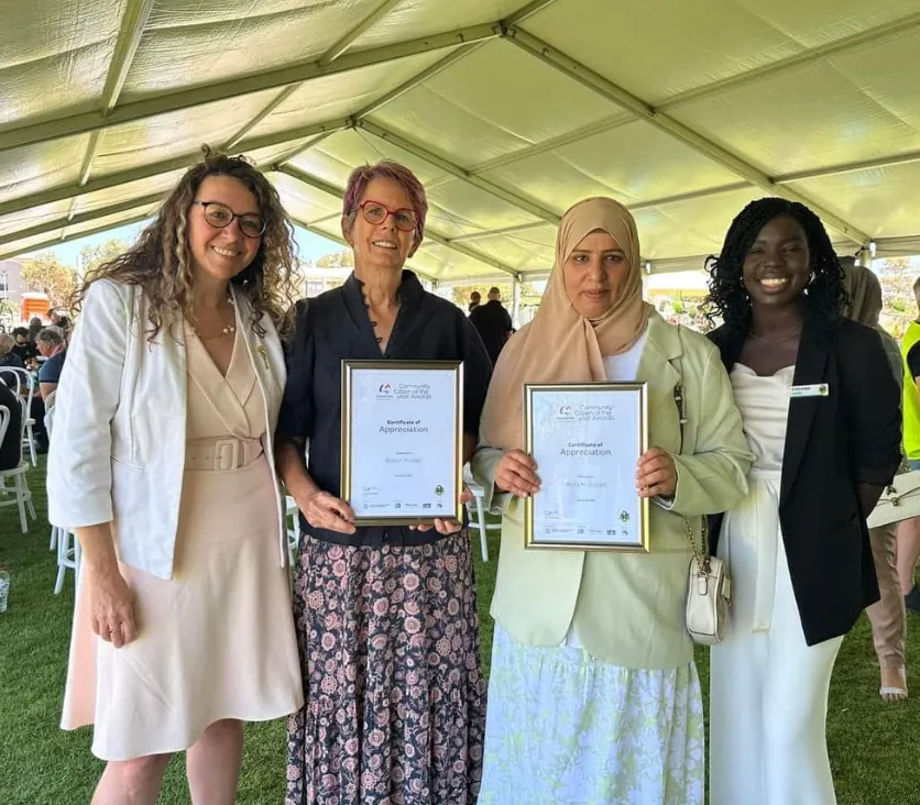 Four women at an outdoor event, two holding framed certificates of appreciation